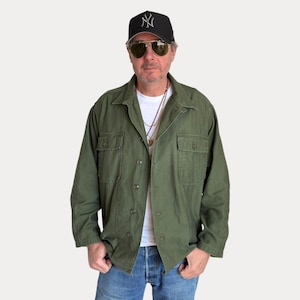 Puede incluir: Una camisa utilitaria de color verde oliva con botones y dos bolsillos en el pecho, sobre una camiseta blanca lisa. El conjunto incluye vaqueros azules, una gorra de béisbol negra con un logotipo blanco y gafas de sol aviador doradas.