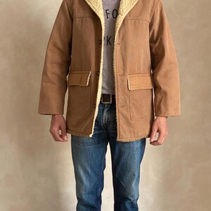 May include: A brown corduroy jacket with a sherpa lining and a brown leather patch on the pocket. The jacket has a button closure and a collar. The person is wearing blue jeans and a grey t-shirt with the word "FOR" on it. The person is also wearing a blue baseball cap with a white logo.