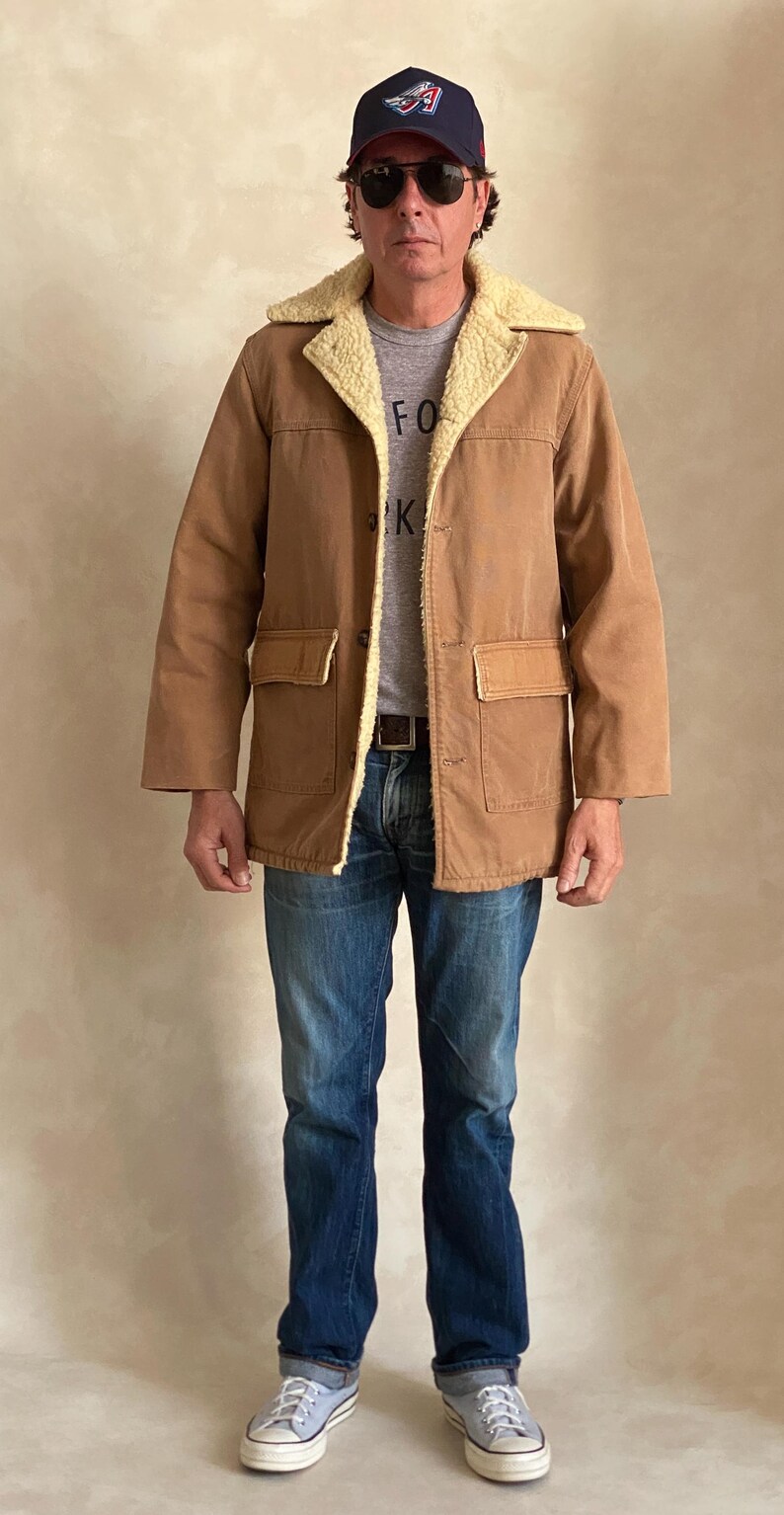 May include: A brown corduroy jacket with a sherpa lining and a brown leather patch on the pocket. The jacket has a button closure and a collar. The person is wearing blue jeans and a grey t-shirt with the word "FOR" on it. The person is also wearing a blue baseball cap with a white logo.