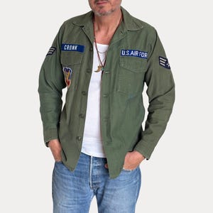 May include: An olive green military-style button-down shirt featuring two chest pockets, a collar, and long sleeves. The shirt displays blue patches with white text reading CRONK and U.S. AIR FORCE, along with additional embroidered insignia patches.