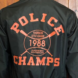 May include: Black baseball jacket with orange and white stripes on the collar and cuffs. The back of the jacket features a large orange graphic that says "Police" with a baseball and bats, "Harrison Little League", "1988", "Eastern Division", and "Champs".