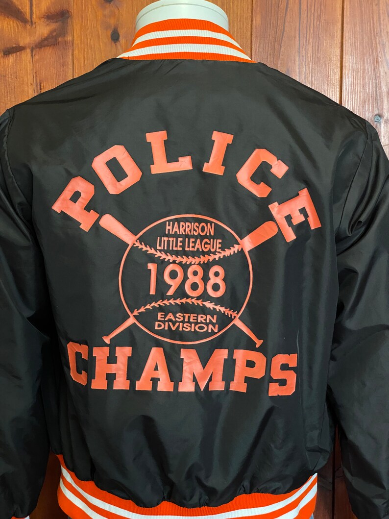 May include: Black baseball jacket with orange and white stripes on the collar and cuffs. The back of the jacket features a large orange graphic that says "Police" with a baseball and bats, "Harrison Little League", "1988", "Eastern Division", and "Champs".