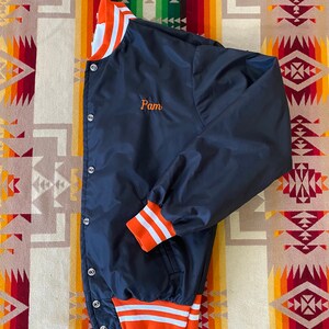May include: A navy blue satin baseball jacket with orange and white striped ribbing at the collar, cuffs, and waistband. The jacket has a button closure and a small embroidered name "Pam" on the left chest.