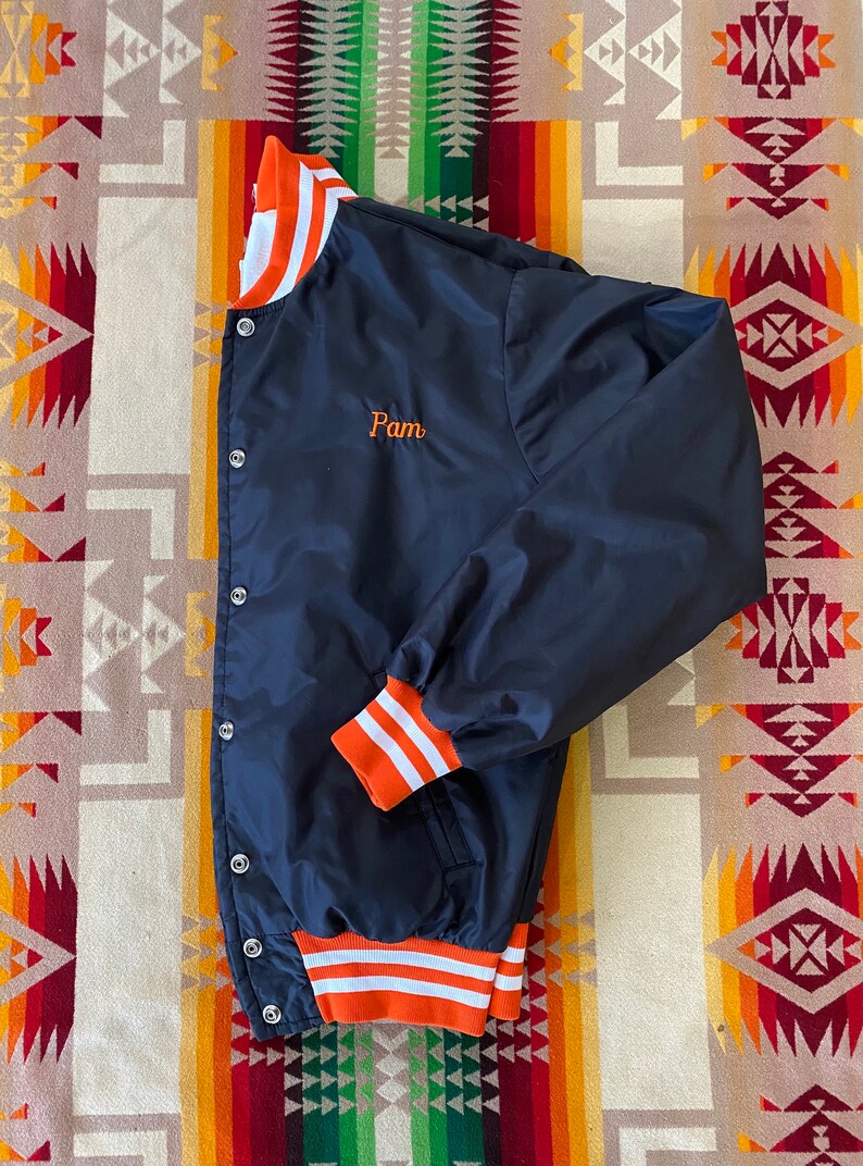 May include: A navy blue satin baseball jacket with orange and white striped ribbing at the collar, cuffs, and waistband. The jacket has a button closure and a small embroidered name "Pam" on the left chest.