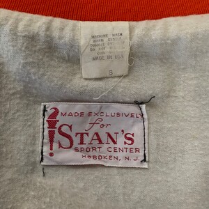 May include: White tag with red text that reads "Made Exclusively for Stan's Sport Center Hoboken, N.J.". A smaller white tag with black text reads "Machine Wash Warm Gentle Tumble Dry Low Do Not Bleach Made in USA S".