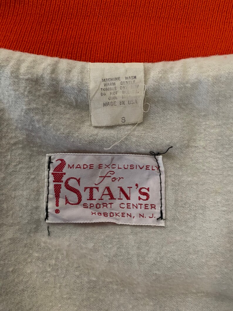 May include: White tag with red text that reads "Made Exclusively for Stan's Sport Center Hoboken, N.J.". A smaller white tag with black text reads "Machine Wash Warm Gentle Tumble Dry Low Do Not Bleach Made in USA S".