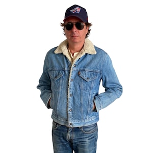 May include: A man wearing a blue denim jacket with a shearling collar, blue jeans, and a navy blue baseball cap with a red, white, and blue logo. He is wearing sunglasses.