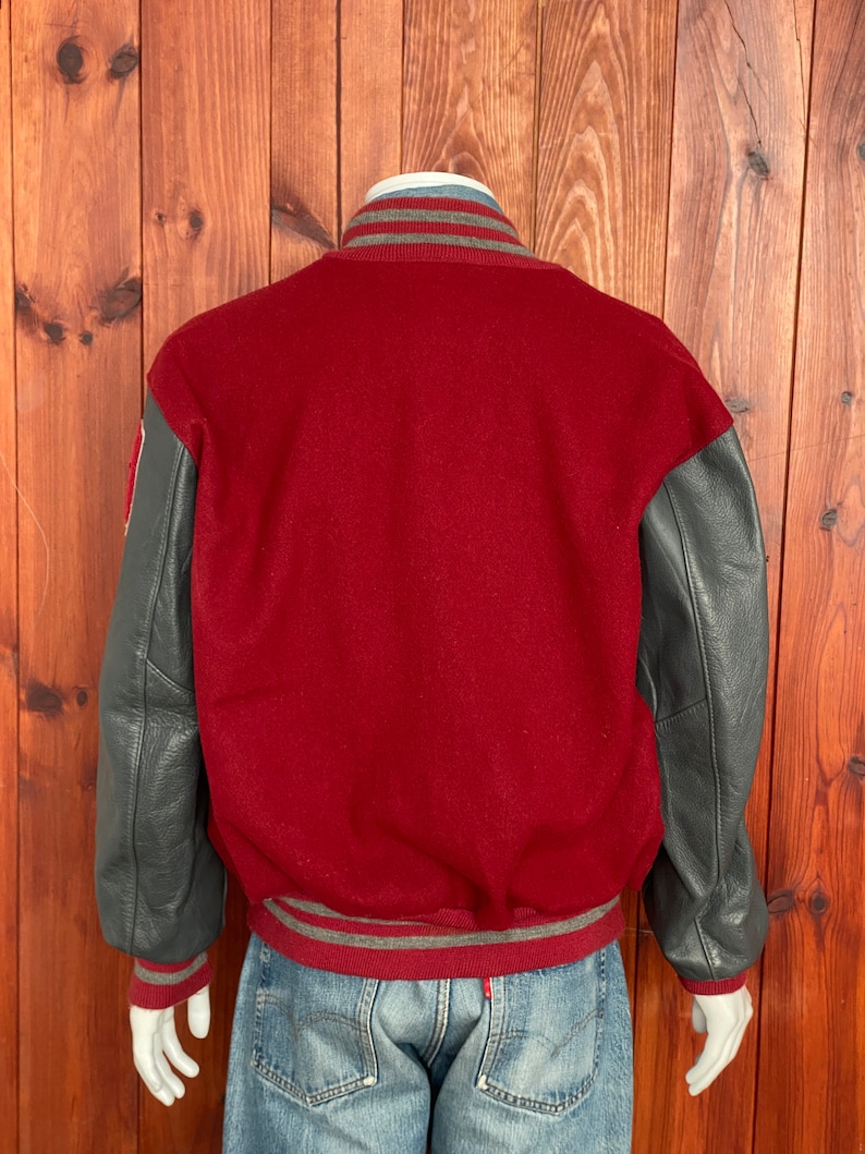 Op de afbeelding: Een bordeauxrode varsity jas met grijze leren mouwen. De jas heeft grijs en wit gestreepte boorden op de kraag, manchetten en taille. De jas wordt op een mannequin getoond.