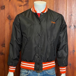 May include: Black satin bomber jacket with orange and white striped ribbing at the collar, cuffs, and hem. The jacket has a button closure and features the word "Pam" embroidered on the left chest in orange.