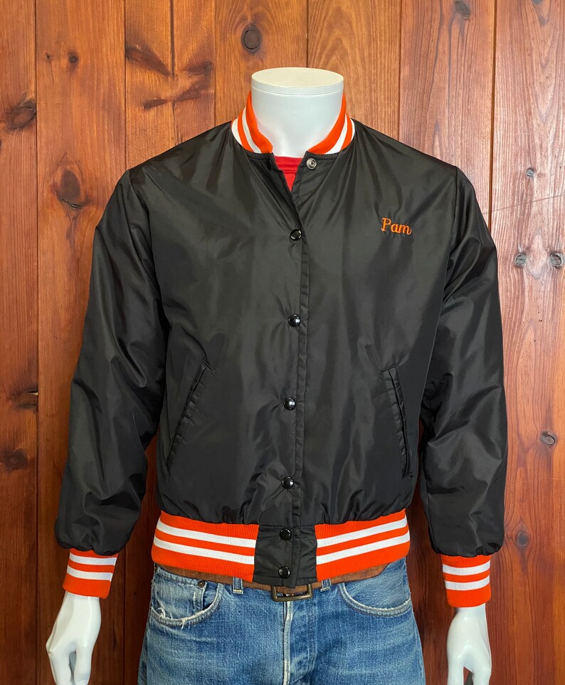 May include: Black satin bomber jacket with orange and white striped ribbing at the collar, cuffs, and hem. The jacket has a button closure and features the word "Pam" embroidered on the left chest in orange.