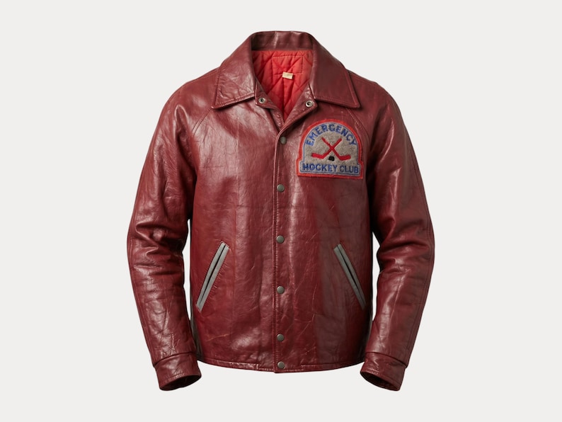 May include: A burgundy leather jacket with a button-up front, a collar, and a red quilted lining. The jacket features a patch on the chest that reads "Emergency Hockey Club" with crossed hockey sticks.