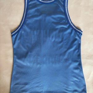 May include: A light blue sleeveless basketball jersey made of a breathable mesh fabric. The garment features a round neckline and armholes trimmed with white and blue striped piping. The overall design is plain with no visible logos or text.