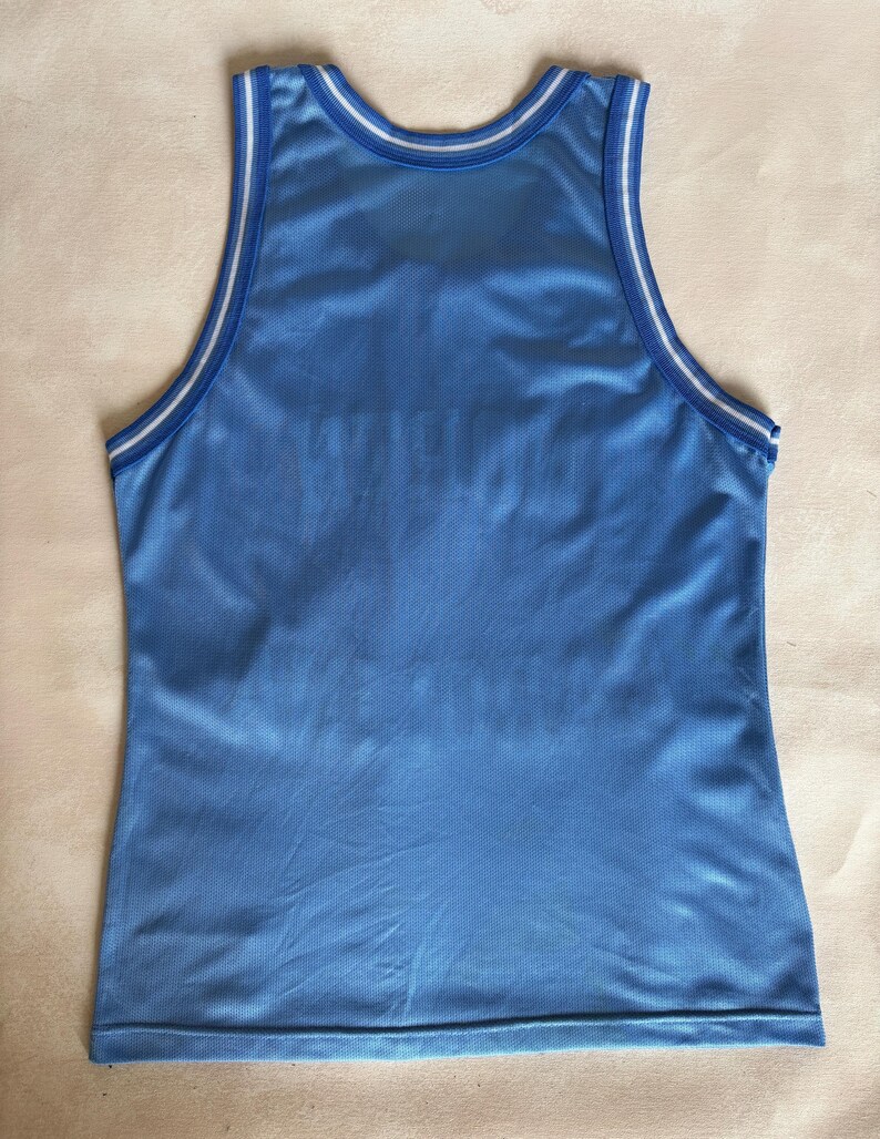 May include: A light blue sleeveless basketball jersey made of a breathable mesh fabric. The garment features a round neckline and armholes trimmed with white and blue striped piping. The overall design is plain with no visible logos or text.