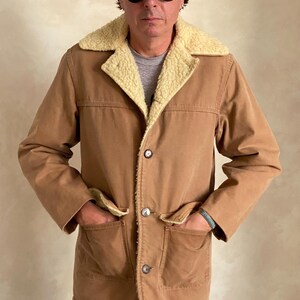 May include: A brown corduroy jacket with a sherpa lining and a button closure. The jacket has two large pockets on the front and a smaller pocket on the chest. The jacket is worn by a person wearing a baseball cap and sunglasses.