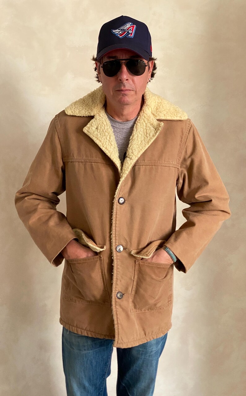 May include: A brown corduroy jacket with a sherpa lining and a button closure. The jacket has two large pockets on the front and a smaller pocket on the chest. The jacket is worn by a person wearing a baseball cap and sunglasses.