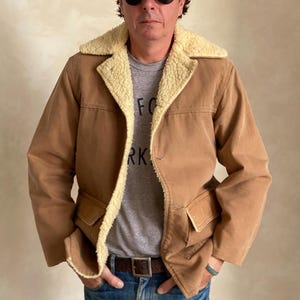 May include: A brown jacket with a sherpa collar and a single button closure. The jacket has two large pockets on the front. The person wearing the jacket is also wearing a grey t-shirt with the word "FOR" and "RK" visible, blue jeans, a brown belt, and a blue baseball cap with a red and white logo.