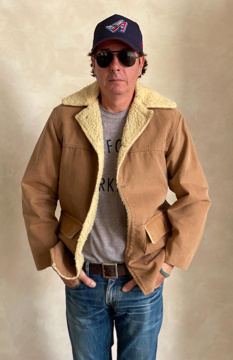 May include: A brown jacket with a sherpa collar and a single button closure. The jacket has two large pockets on the front. The person wearing the jacket is also wearing a grey t-shirt with the word "FOR" and "RK" visible, blue jeans, a brown belt, and a blue baseball cap with a red and white logo.