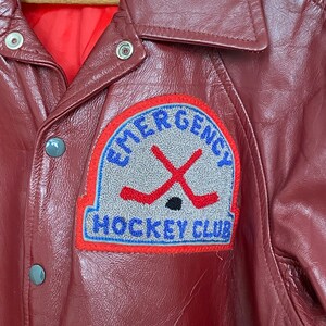 May include: A burgundy leather jacket with a red quilted lining. The jacket features a patch with the words "EMERGENCY HOCKEY CLUB" and crossed hockey sticks. The jacket has a button-up front and a collared neckline.