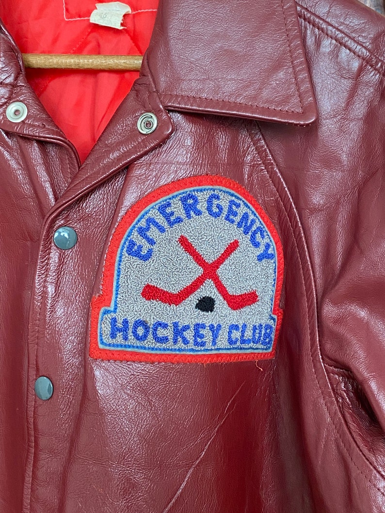 May include: A burgundy leather jacket with a red quilted lining. The jacket features a patch with the words "EMERGENCY HOCKEY CLUB" and crossed hockey sticks. The jacket has a button-up front and a collared neckline.