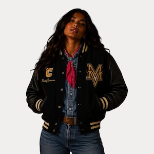 May include: Black varsity jacket with gold and white detailing. The jacket has a large "M" and "C" patch with text. It features a button-up front, black leather sleeves, and striped cuffs and hem. A pink scarf is worn around the neck.