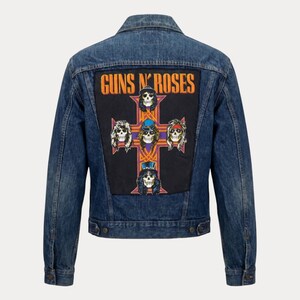 May include: A blue denim jacket with a Guns N' Roses patch on the back. The patch features the band's name in orange and a cross design with skull illustrations. The jacket has a classic button-up design with pockets and long sleeves.