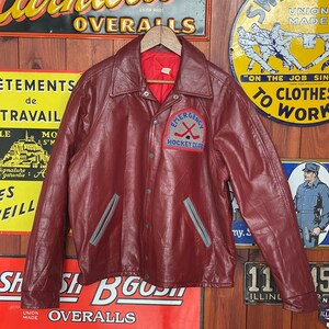 May include: A burgundy leather jacket with a button-up front and a collar. The jacket features gray accents and a patch that reads "EMERGENCY HOCKEY CLUB." The jacket is hanging on a wooden hanger against a backdrop of vintage signs.