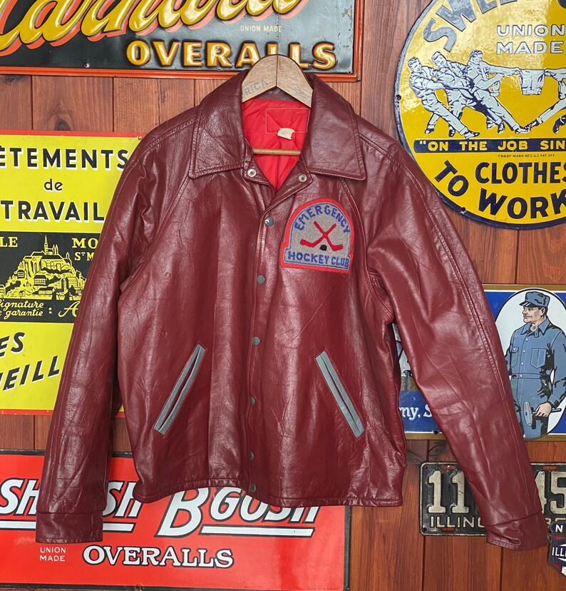 May include: A burgundy leather jacket with a button-up front and a collar. The jacket features gray accents and a patch that reads "EMERGENCY HOCKEY CLUB." The jacket is hanging on a wooden hanger against a backdrop of vintage signs.