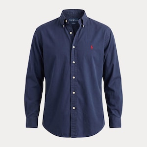 May include: A navy blue button-down shirt with long sleeves. The shirt features a classic collar and a small red logo on the chest. The shirt has white buttons and is made of a soft, woven fabric.