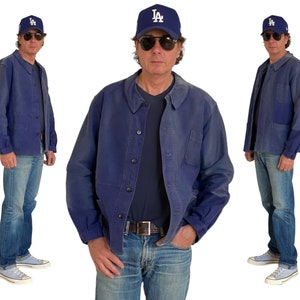May include: A man wearing a blue denim jacket with a blue baseball cap that says "LA" on it. He is also wearing blue jeans and white sneakers.