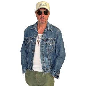 May include: A denim jacket with a light wash, worn over a white undershirt and olive green pants. The person is wearing a cream-colored baseball cap with "Mets" embroidered on the front and aviator sunglasses.