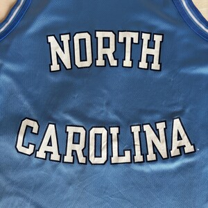 May include: A light blue sleeveless basketball jersey featuring the text NORTH CAROLINA in white letters with dark blue outlines. The jersey has a white and dark blue striped collar and a Champion logo patch on the upper left chest area.