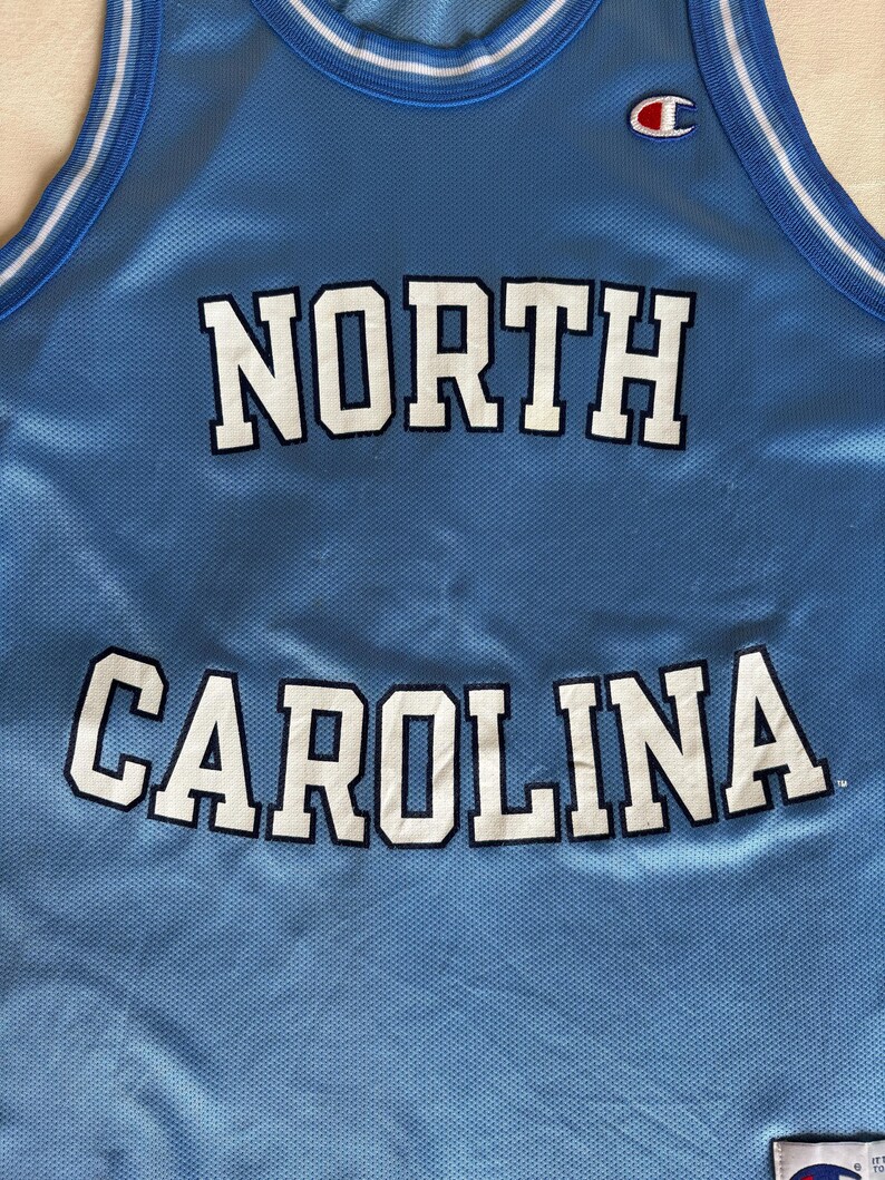 May include: A light blue sleeveless basketball jersey featuring the text NORTH CAROLINA in white letters with dark blue outlines. The jersey has a white and dark blue striped collar and a Champion logo patch on the upper left chest area.