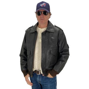 Puede incluir: Una chaqueta bomber de cuero marrón con cierre de cremallera y dos bolsillos de solapa. La chaqueta tiene un cuello, puños y cintura de punto marrón. El modelo lleva una camiseta blanca y unos vaqueros azules.