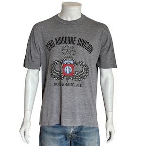 May include: Gray t-shirt featuring the text "82ND AIRBORNE DIVISION" above a military emblem with wings, a star, and the letters "AP" inside a red and blue shield. The text "FOR BRAGG, N.C." is printed below the emblem.