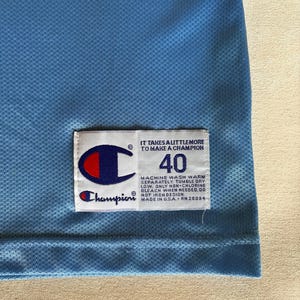 May include: A light blue mesh jersey featuring a white rectangular tag with the Champion logo, the number 40, and the text: IT TAKES A LITTLE MORE TO MAKE A CHAMPION. MACHINE WASH WARM SEPARATELY. TUMBLE DRY LOW. ONLY NON-CHLORINE BLEACH WHEN NEEDED. DO NOT IRON DESIGN. MADE IN U.S.A. RN 26094.