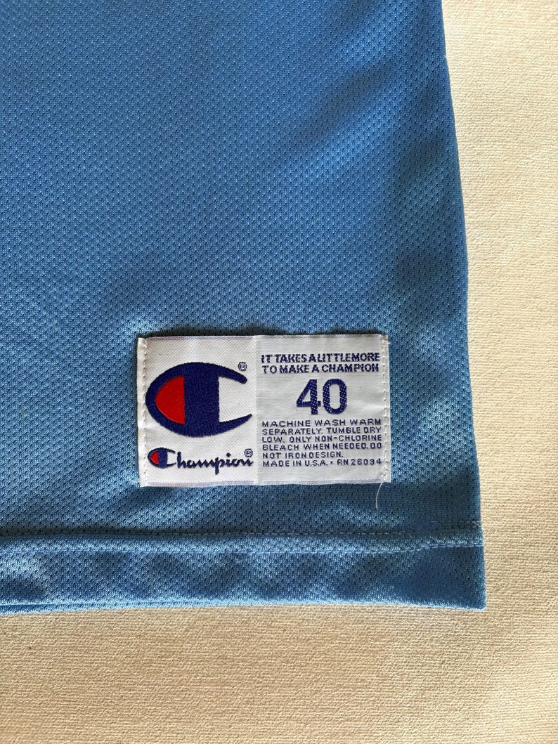 May include: A light blue mesh jersey featuring a white rectangular tag with the Champion logo, the number 40, and the text: IT TAKES A LITTLE MORE TO MAKE A CHAMPION. MACHINE WASH WARM SEPARATELY. TUMBLE DRY LOW. ONLY NON-CHLORINE BLEACH WHEN NEEDED. DO NOT IRON DESIGN. MADE IN U.S.A. RN 26094.