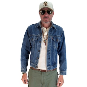 May include: A denim jacket with button closures, worn over a cream-colored shirt. The person is wearing a baseball cap with a logo, sunglasses, and a necklace. The outfit includes green cargo pants and a brown belt.