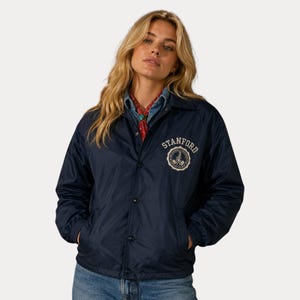 May include: A navy blue coach jacket with the word "STANFORD" and a circular emblem on the chest. The jacket has a button-up front and pockets. The person is wearing blue jeans.