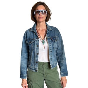 May include: A medium-wash denim jacket with a collared neckline and button-up front. The jacket is paired with a white top and olive green cargo pants. The person is wearing white sunglasses and a necklace.