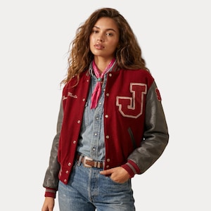 Op de afbeelding: Een rode varsity jas met grijze mouwen en een groot wit "J"-embleem. De jas heeft een knoopsluiting en de naam "Martin" geborduurd op de borst. Gedragen met een spijkerblouse en een blauwe jeans.