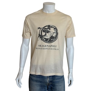 Könnte beinhalten: Ein cremefarbenes T-Shirt mit einer kreisförmigen Grafik mit Biathleten. Die Grafik ist in Schwarz und Grau gehalten, darunter steht der Text "SKAGENAPOLI TRANS-EUROPEAN BIATHLON". Das Shirt wird auf einer Schaufensterpuppe präsentiert.