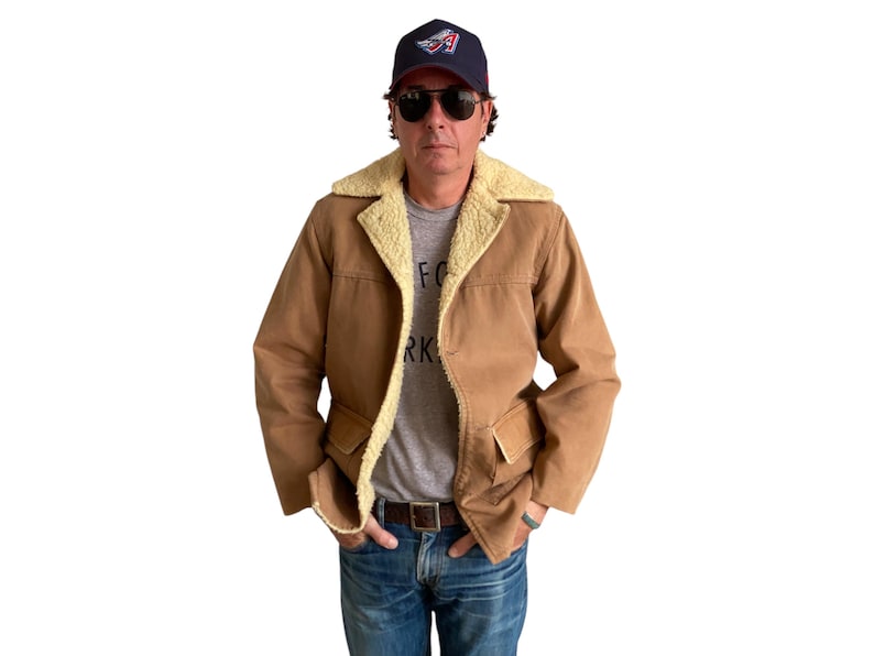 May include: A brown suede jacket with a shearling collar and two front pockets. The jacket is worn by a person wearing a grey t-shirt with the text "FOR NEW YORK" printed on it, blue jeans, and a blue baseball cap with a red, white, and blue logo.