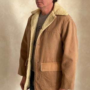 May include: A brown corduroy jacket with a sherpa lining. The jacket has a button closure and a large pocket on the front. The person wearing the jacket is also wearing a blue baseball cap with a red brim and a white logo.