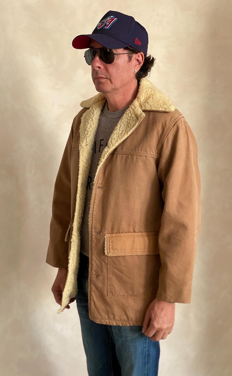 May include: A brown corduroy jacket with a sherpa lining. The jacket has a button closure and a large pocket on the front. The person wearing the jacket is also wearing a blue baseball cap with a red brim and a white logo.