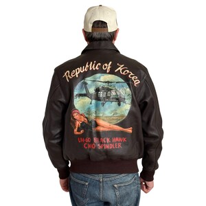 Puede incluir: Chaqueta de cuero marrón con un gráfico detallado en la espalda. El gráfico presenta un helicóptero, una mujer y el texto "Republic of Korea" y "UH-60 Black Hawk CWO Spindler".
