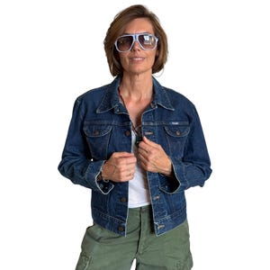 May include: A dark blue denim jacket with a button-up front, two chest pockets, and a collar. The jacket has a Wrangler logo patch. The person is wearing light blue sunglasses and green cargo trousers. The background is white.