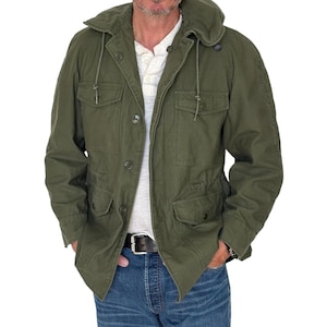 May include: An olive green field jacket featuring a hood with drawstring cords, a front zipper closure with buttoned storm flap, two chest pockets with buttoned flaps, and two lower pockets with buttoned flaps, worn over a white shirt and blue jeans.