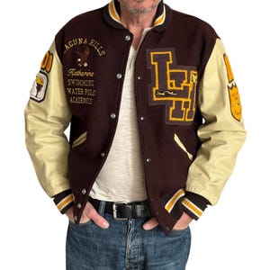 Op de afbeelding: Een bruine varsity-jas met crèmekleurige mouwen, voorzien van de tekst Laguna Hills, Katherine, Swimming, Water Polo Academy en de letters LH. De jas heeft een drukknoopsluiting aan de voorzijde en geribbelde boorden met gele strepen.