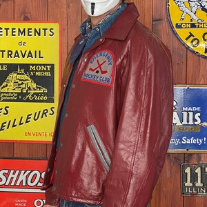 May include: A burgundy leather jacket with a gray stripe detail and a patch that reads "Emergency Hockey Club." The jacket has a button-up front and a collared neckline. The person is wearing a black bowler hat and a white mask.