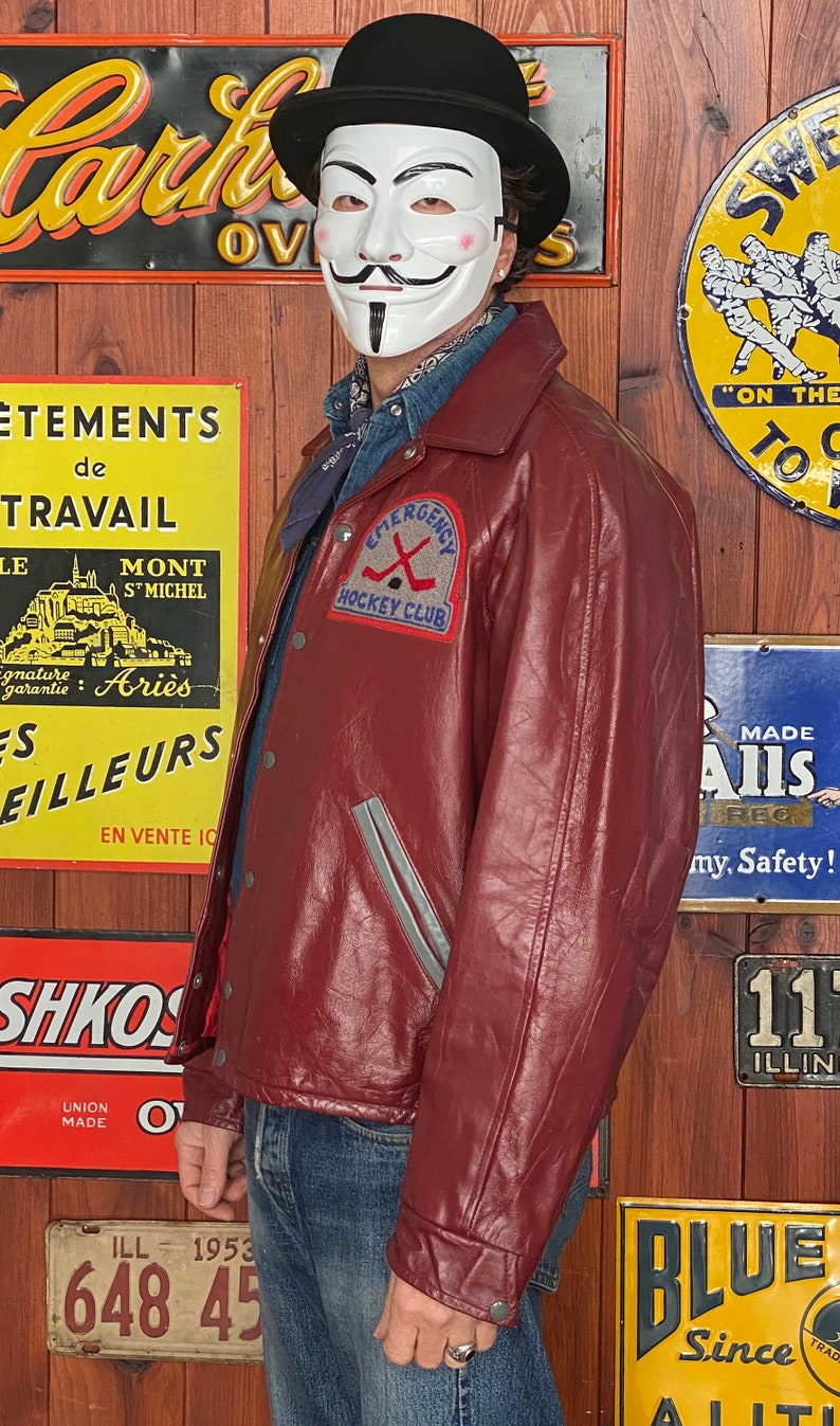 May include: A burgundy leather jacket with a gray stripe detail and a patch that reads "Emergency Hockey Club." The jacket has a button-up front and a collared neckline. The person is wearing a black bowler hat and a white mask.